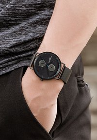 Orologio analogico nero con due sottodial su un quadrante nero testurizzato. Presenta un cinturino in rete metallica e una cassa rotonda minimalista.