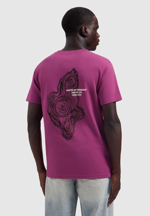 Man wearing purple t-shirt with black tree ring graphic and text "Rooted by friendship, tree of life, pure path" on the back.