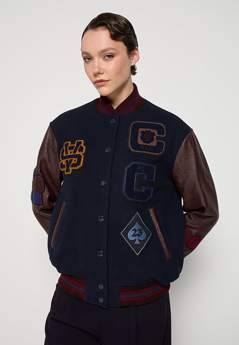Giovane con una giacca varsity blu navy con maniche in pelle marrone e vari toppe ricamate colorate, in piedi davanti a uno sfondo bianco.