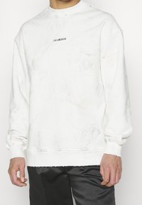 Bílá mikina s texturovaným vzorem, žebrovaným kulatým výstřihem a manžetami. Na přední části uprostřed je malá černá značka.