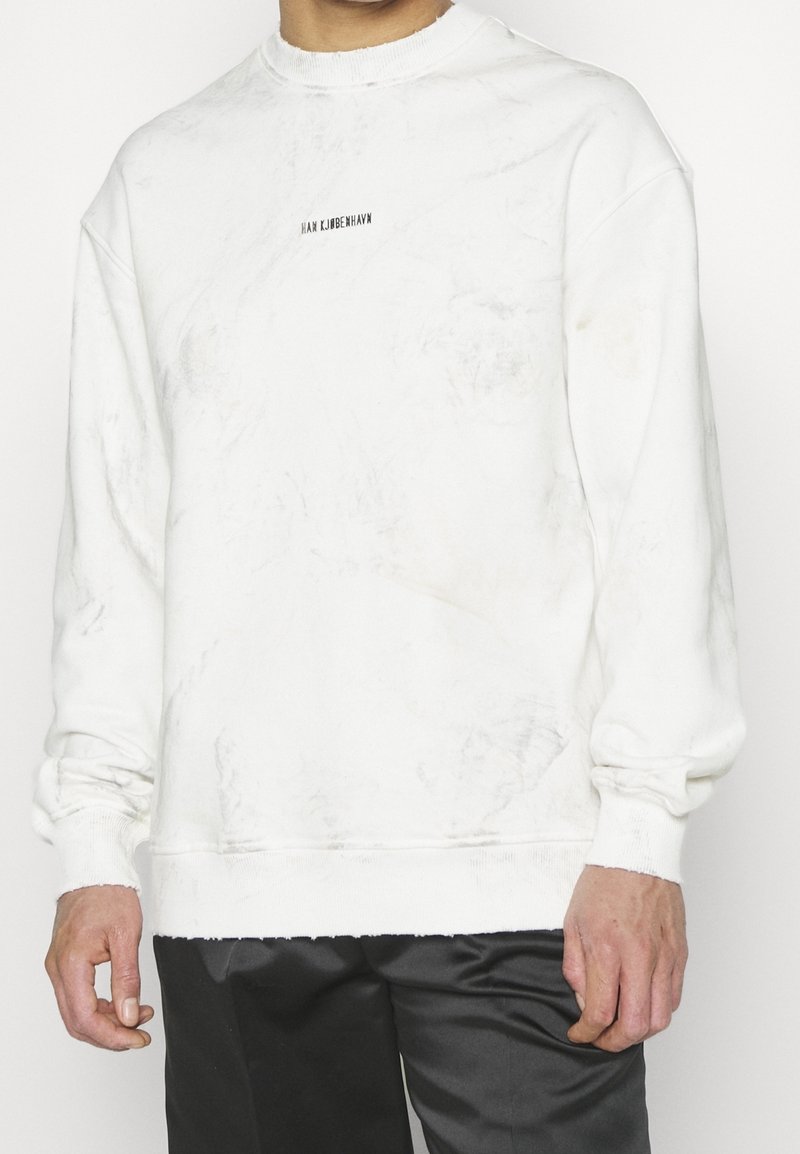 Bílá mikina s texturovaným vzorem, žebrovaným kulatým výstřihem a manžetami. Na přední části uprostřed je malá černá značka.