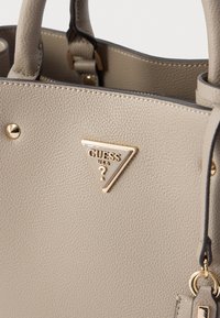 Beigefarbene Handtasche aus strukturiertem Leder mit goldfarbenen Beschlägen, einschließlich einer dreieckigen Guess USA Logo-Plakette und dekorativen Nieten.