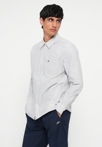 Striped button-up shirt in white and blue, featuring a small green logo on the chest pocket. Worn with navy trousers.