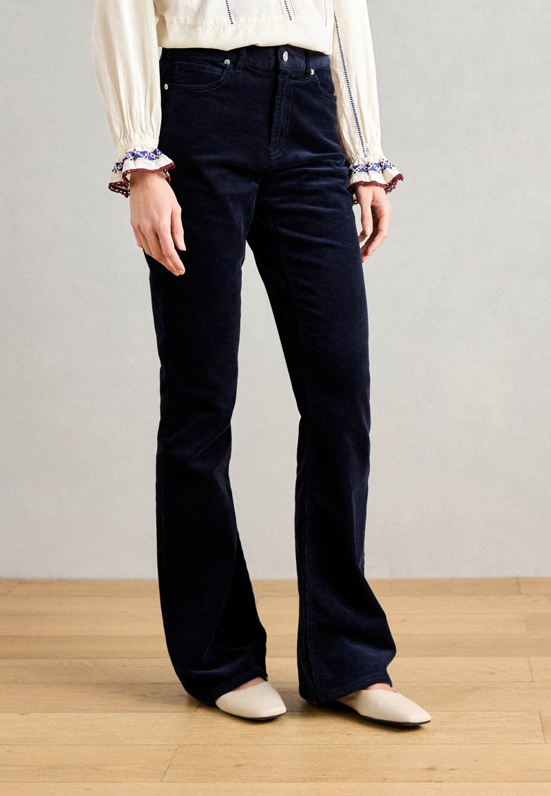 Navyblaue Cordhosen mit weitem Bein, ausgestattet mit vier Taschen, hoher Taille und glatter Textur. Kombiniert mit cremefarbenen Slippern.