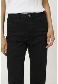 Personne portant un jean noir taille haute et un haut blanc sans manches, debout avec les bras détendus le long du corps.