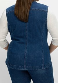 Denimväst i mörkblått med en figursydd design, med sömmar längs ryggen och rundad nederkant; tillsammans med en långärmad vit skjorta.