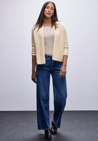 Un cárdigan texturizado en crema sobre una blusa beige, combinado con unos pantalones vaqueros de pernera ancha en azul y botas negras hasta el tobillo, sobre un fondo neutro.