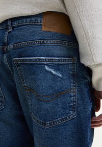Jeans in denim blu scuro con una texture invecchiata, dotati di un'etichetta in pelle marrone e cuciture visibili sulle tasche posteriori.