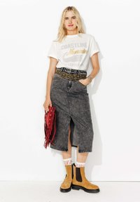 T-shirt blanc graphique avec le texte "COASTLINE Memories", associé à une jupe midi en denim foncé avec une fente avant et une ceinture décorative. Sac frangé bordeaux et bottines beiges avec semelles noires.