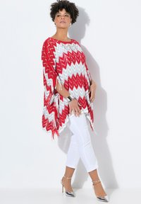 Poncho a zigzag rosso e bianco con dettagli in frangia, abbinato a pantaloni bianchi aderenti e tacchi alti argentati. Texture liscia, vestibilità morbida.