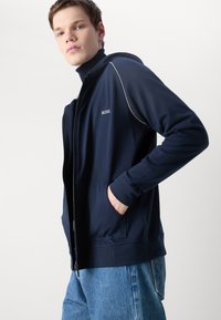 Felpa con zip navy, con cuciture e logo bianchi. Presenta una tasca frontale, una vestibilità comoda e una texture morbida. Abbinata a jeans chiari.