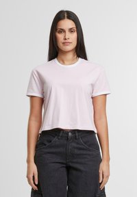 Lys rosa cropped t-skjorte med korte ermer og hvit ribbestrikket hals, laget av glatt stoff. Brukt med høyt liv svarte bukser.