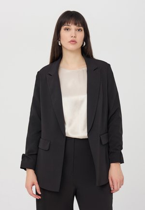 Blazer nero con un design sartoriale, due tasche frontali e una texture liscia, sovrapposto a un top leggero e a coste. Estetica minimalista.