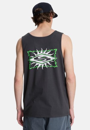 Homme portant un débardeur noir avec un motif de soleil vert et blanc, une casquette sombre portée à l'envers, et un short bleu, debout de dos à la caméra.