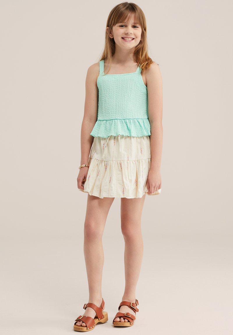 Fille souriante, portant un haut sans manches turquoise clair avec un ourlet à volants, une jupe à étages crème à motifs, des sandales marron à boucle et un bracelet.