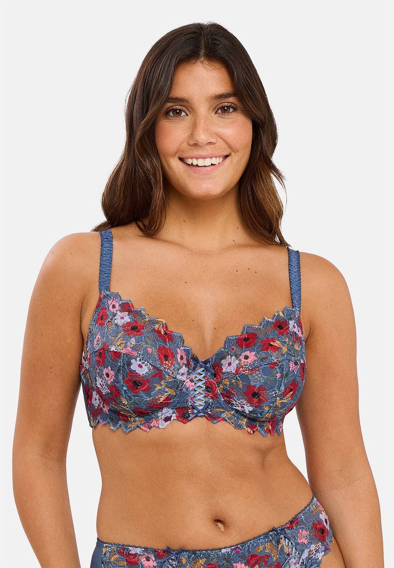 Lachende vrouw die een bijpassende blauwe kanten bh en slipje draagt met rode, roze en gele bloemen op een effen achtergrond.