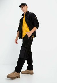 Sort fleecejakke over en gul skjorte, parret med sorte bukser med sidelomme og brune støvler, sat mod en hvid baggrund.