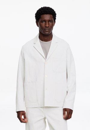 Mężczyzna w białej zapiętej kurtce na jasnoszarej koszuli, stojący na jednolitym białym tle.