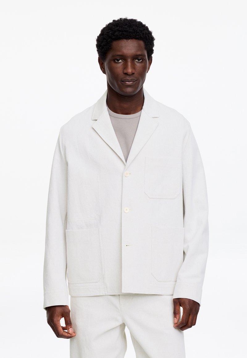Homme portant une veste blanche boutonnée sur une chemise gris clair, debout devant un fond blanc uni.