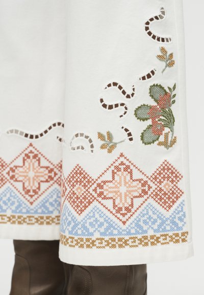 Λευκό ύφασμα με ορατά καφέ μποτάκια, διακοσμημένο με κεντητά γεωμετρικά σχέδια σε καφέ, μπλε και ανθικά μοτίβα, με λεπτομέρειες κοψίματος κοντά στο τελείωμα.