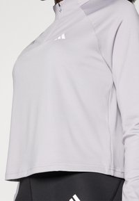 Vaaleanharmaa pitkähihaurheilupaita, jossa on vetoketjullinen pääntie ja kolme valkoista raitaa edessä. Paita on väljää istuvuutta ja sileää pintaa.