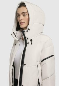 Hvid pufferjakke med hætte, der har sorte lynlåse og detaljer. Materialet ser glat ud med en quilted tekstur.