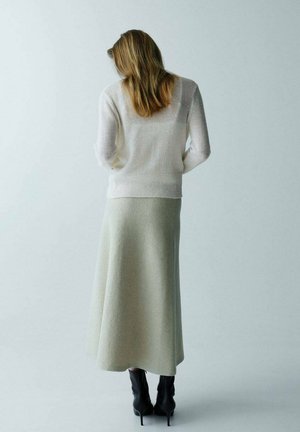 Mujer con cabello castaño claro, vestida con un suéter blanco, falda midi beige y botas negras de tacón, de pie frente a un fondo claro y liso.