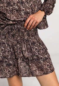 Robe en tissu à motifs bruns avec une taille smockée, des volants à plusieurs niveaux et des manches longues. Le matériau semble léger et texturé.