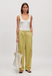 Lichtgele cargobroek met zijzakken, gecombineerd met een witte, getailleerde tanktop. Een crèmekleurige schoudertas wordt in één hand vastgehouden.