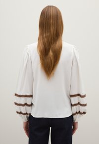 Blusa blanca con mangas largas y abullonadas que presenta bordados en zigzag marrón cerca de los puños. Textura suave, detalle de botones en los extremos de las mangas.