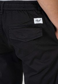 Pantalones negros con un bolsillo trasero único con tapa en ángulo, cinturilla elástica y una etiqueta blanca con el texto "Reell est. 1997."
