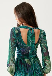 Žena s dlouhými tmavými vlasy nosí zelené šaty s tropickým vzorem listů, které mají dlouhé průsvitné rukávy a otevřený zadní díl zavázaný na krku.