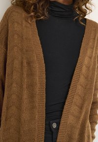 Brauner gestrickter Cardigan mit einem strukturierten Zickzackmuster, offener Vorderseite und gerippten Kanten, über einem schwarzen Rollkragenoberteil getragen.