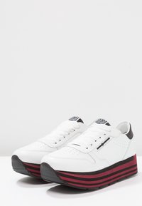 Sneakers bianchi con tomaia in pelle liscia, dettagli neri sul tallone e una suola piatta stratificata con strisce orizzontali rosse e nere.
