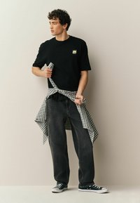 Fiatal férfi fekete grafikás pólóban, fekete farmerban, tornacipőben, fekete-fehér kockás inget kötve a derekán, egyszínű fal előtt.