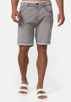 Homme portant un short en denim gris retroussé et des sandales blanches à boucle, debout devant un fond blanc uni.