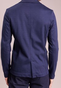 Námořnická modrá saková bunda na míru s dlouhými rukávy, strukturovaná tkanina, zadní rozparek a jemné švy, které podtrhují přiléhavý siluetu.