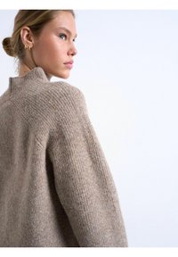 Femme aux cheveux blonds en chignon, portant des boucles d'oreilles créoles dorées et un pull en laine beige texturé, légèrement de profil sur un fond blanc uni.
