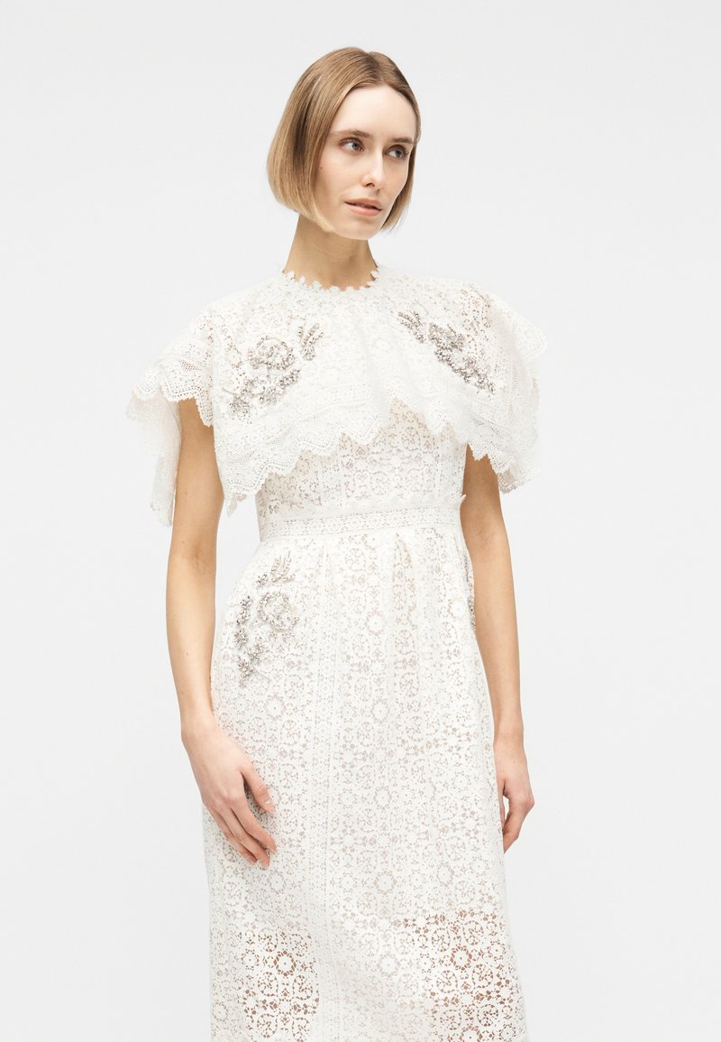 Frau in weißem Spitzenkleid mit floraler Stickerei und kapuzenähnlichem Kragen, stehend vor einfachem hellem Hintergrund.