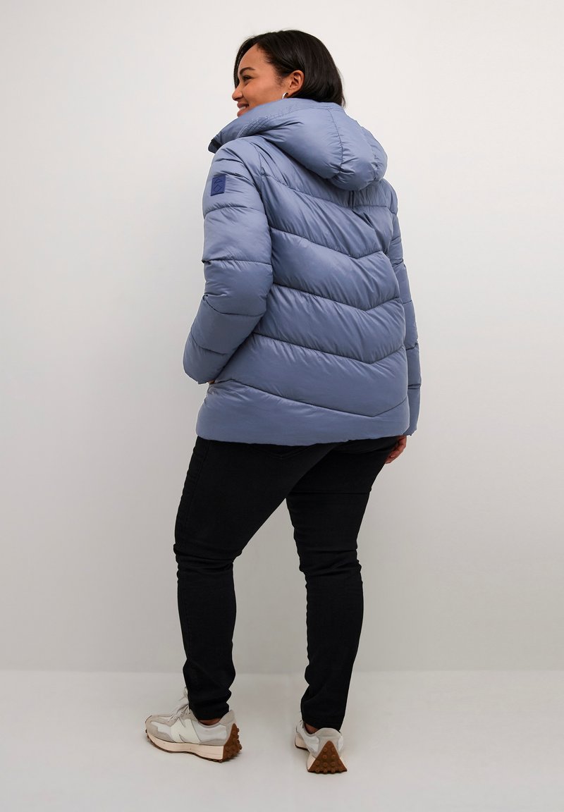 Veste rembourrée bleu clair avec capuche, présentant une texture matelassée horizontale et une coupe légèrement cintrée. Portée sur un jean noir et des baskets.