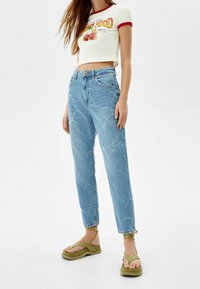 Ljusblå högmidjade jeans med hjärtformad sömnad, parat med en kort vit topp med röda accenter och gröna plattformssandaler.