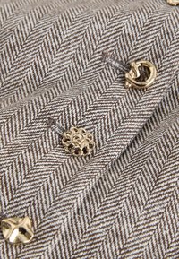 Țesătură în model pepită maro și alb, cu trei nasturi strălucitori de culoare auriu, unul rotund, unul răsucit și unul cu formă neregulată.