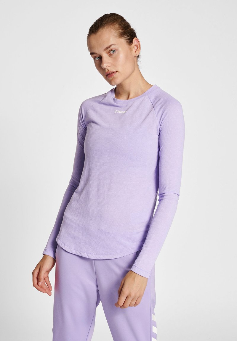 Vrouw draagt lichtpaarse top met lange mouwen en bijpassende broek, staand tegen een effen witte achtergrond.