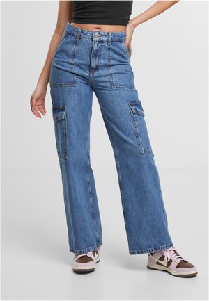 Kvinde iført blå højtaljede vidde cargobukser i denim med lommer foran og på siden, kombineret med pink og brune sneakers samt en sort crop top.