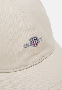 Beige baseball caps laget av teksturert stoff med et sydd logo med røde og blå detaljer. Buet skjerm og synlige sømmer.
