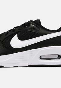 Musta lenkkari, jossa on teksturoitu nahka- ja verkkopinta, valkoisella Nike-logolla, kontrastoivalla valkoisella välipohjalla ja ilmavälityksellä.