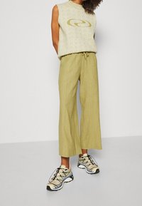 Ljus olivgrön ärmlös topp med en slinga-logotyp, matchad med lösa, strukturerade olivgröna byxor och beigea sportskor med blå accenter.