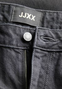 Κοντινή λήψη στην μέση μαύρου τζιν με ετικέτα από ύφασμα JJXX, μεταλλικό κουμπί με χαραγμένο λογότυπο και μερικώς ανοιχτό φερμουάρ.