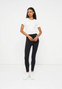 Svarta högmidjade leggings med en slät yta och logotypdetalj, tillsammans med en åtsittande vit t-shirt med kort ärm. Modellen står framför en neutral bakgrund.