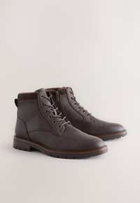 Bottes cheville en cuir marron avec col texturé, lacets à l'avant, fermeture éclair latérale et semelle en caoutchouc avec relief antidérapant. Équipées d'œillets métalliques.